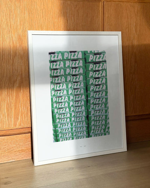 Framed modern art print featuring a stacked pattern of pizza boxes, leaning against a wooden wall